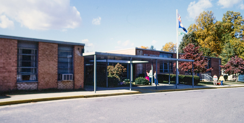 Color photograph of the exterior of West Springfield Elementary school from a 35 millimeter slide. The date the photograph was taken is unknown, but is thought to be in the late 1980s based on the clothing of the people in the image. The photograph shows the front of the building, including the main entrance, and was taken during the fall because the leaves on the trees have started to change color. 