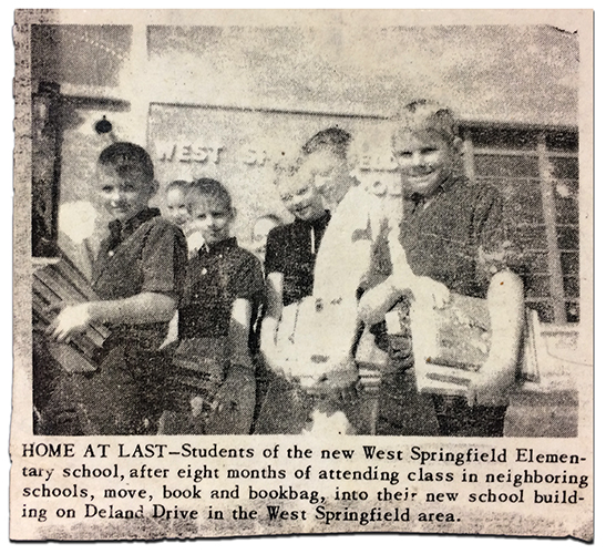 Newspaper article photograph with caption showing seven smiling children in front of West Springfield Elementary School. The children closest to the camera, all boys, are carrying armfuls of books. The image caption reads: Home at Last – Students of the new West Springfield Elementary School, after eight months of attending class in neighboring schools, move, book and backbag, into their new school building on Deland Drive in the West Springfield area. 